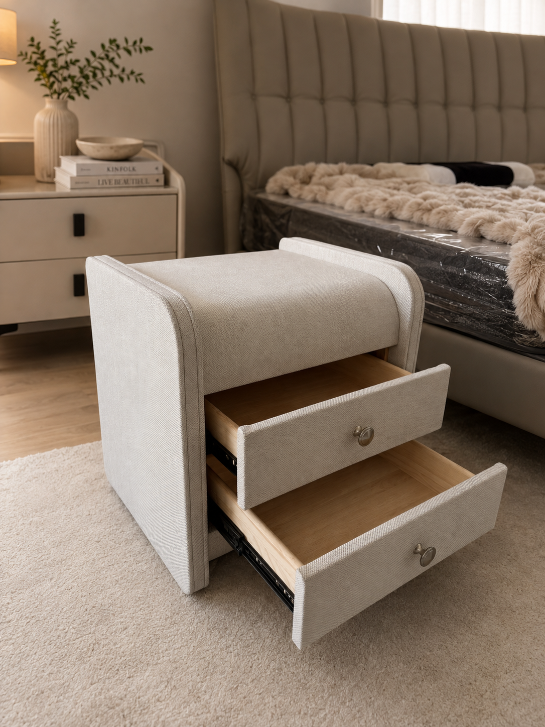 A light beige upholstered bedside table with two open drawers, shown in a modern bedroom setting next to a bed available at Duvisions Casa showrooms in Melbourne and Sydney. 