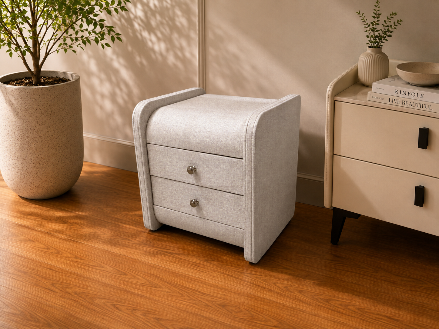A light gray upholstered two-drawer nightstand with rounded edges and small silver knobs, shown in a styled room setting with a wooden floor, potted plant, and cream-colored side table available at Duvisions Casa in Melbourne and Sydney. 